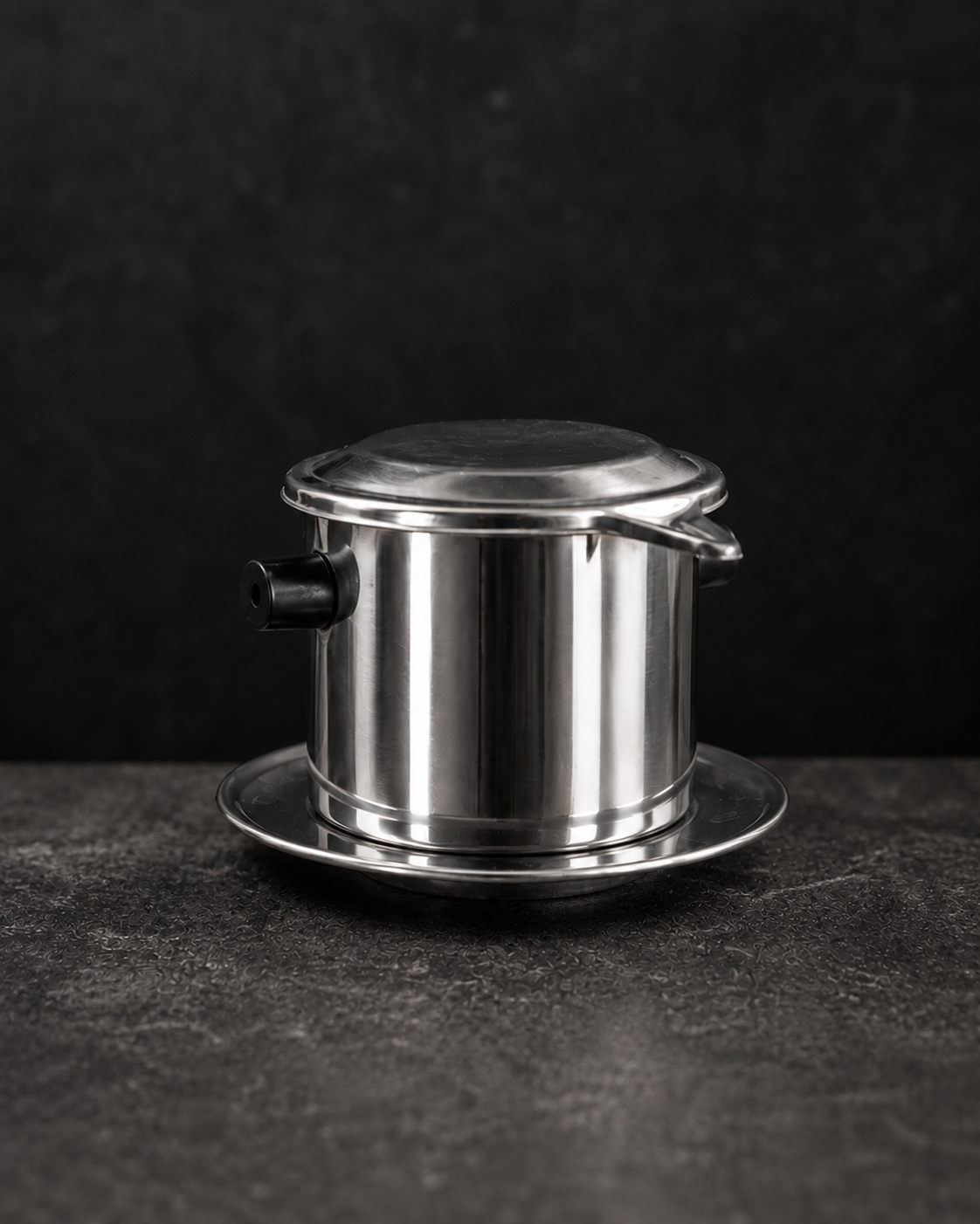 A stainless steel Vietnam-style coffee dripper (phin) with a matching lid and saucer, set against a dark textured background.