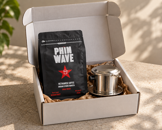 A coffee gift set in an open white cardboard box, featuring a black bag of Phin Wave Vietnamese Coffee and a stainless steel pour-over dripper, displayed on a stone surface with soft natural shadows.